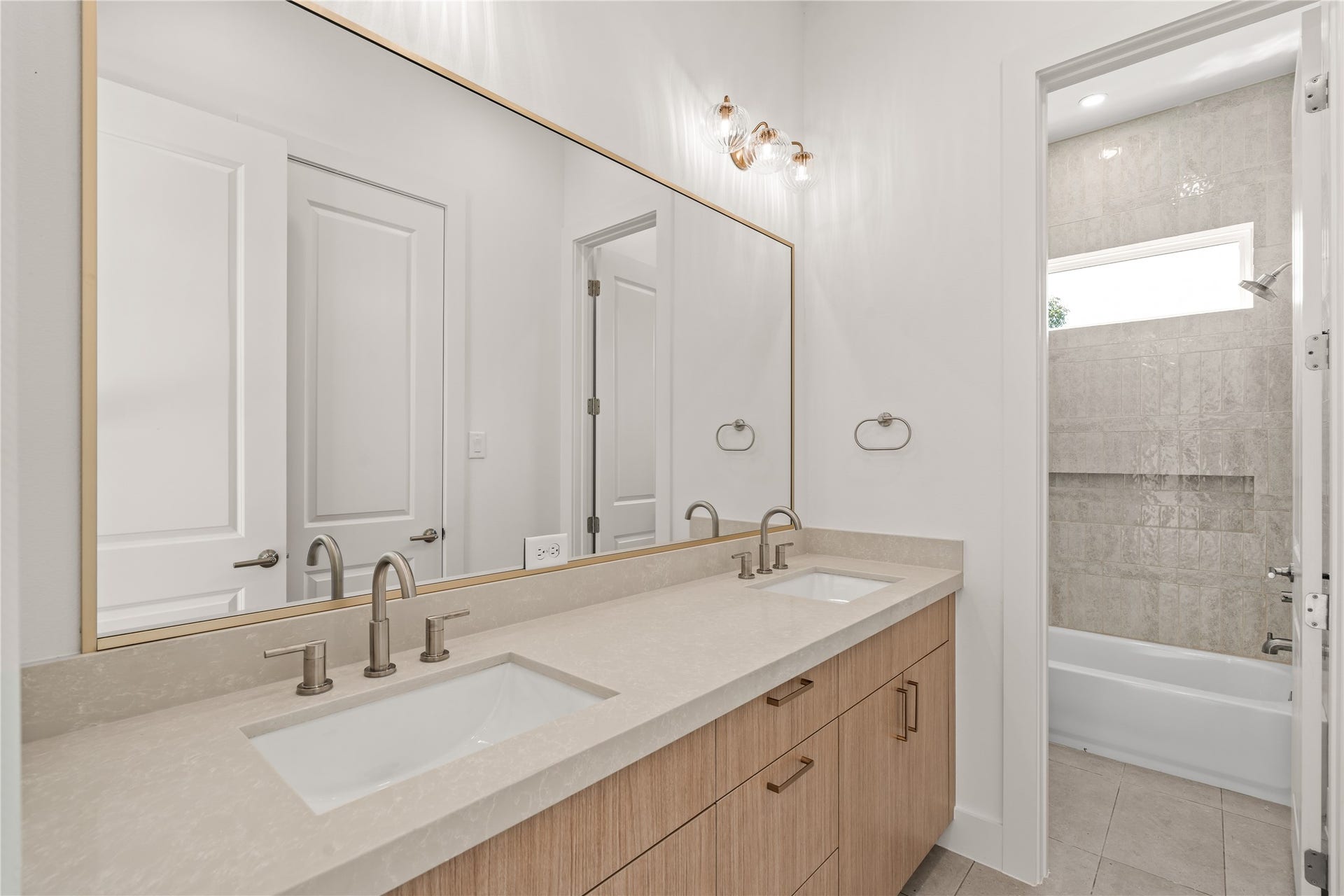 The secondary bath. Double sinks under one wide gold-framed mirror, a wood vanity in quartz, and a tiled tub and shower combo behind the door.