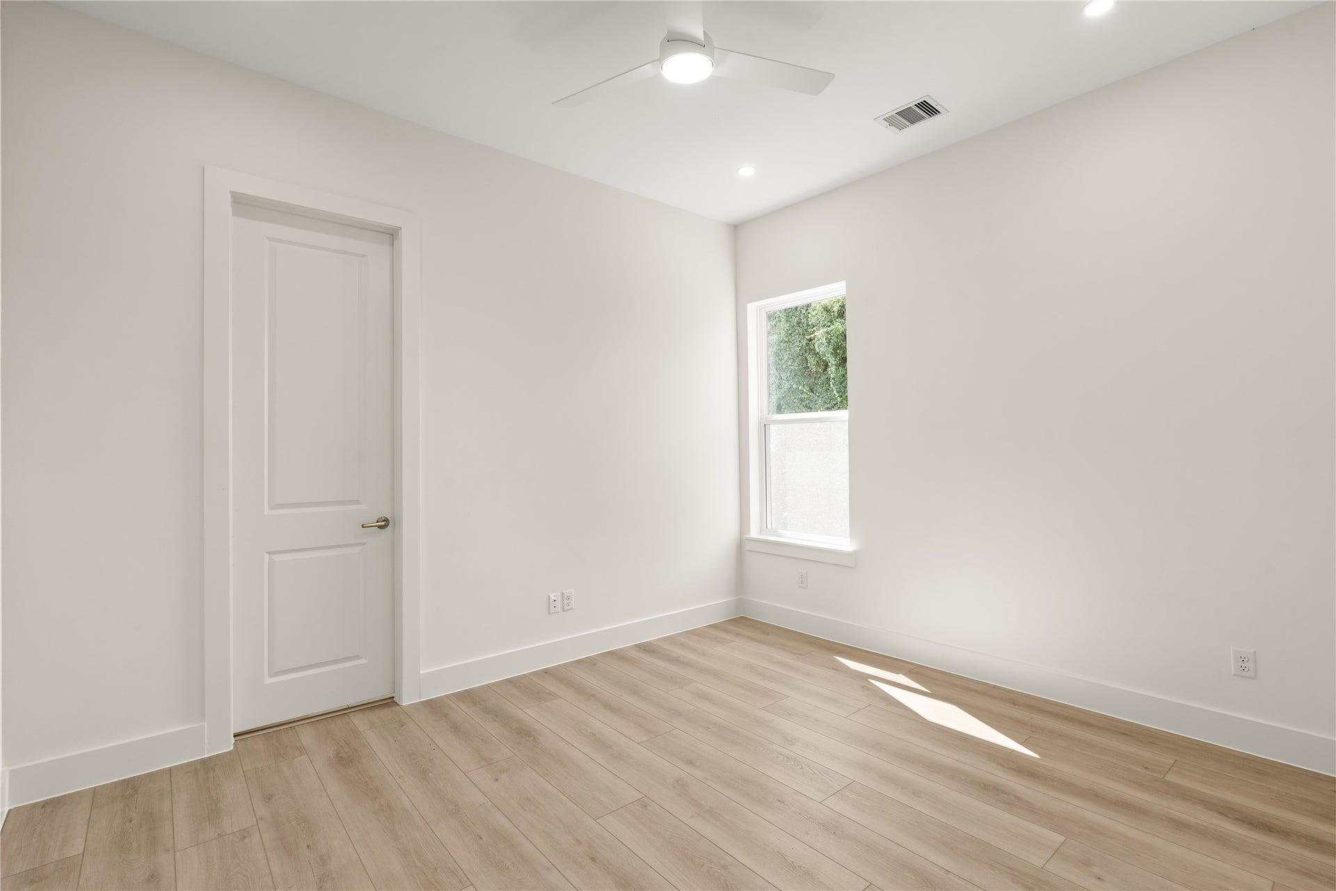 Upstairs bedroom with a tree-shaded window and the closet behind the door. Light wood floors, ceiling fan, no carpet anywhere.