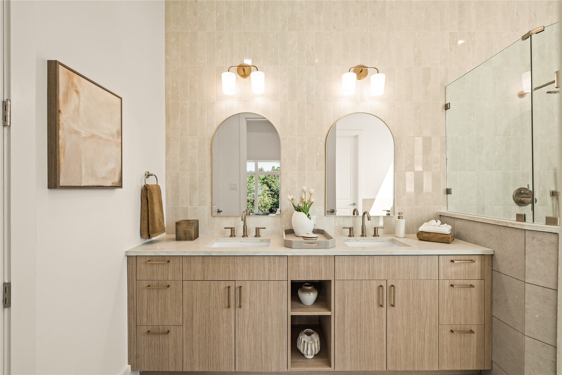 Primary bath. Double sinks under arched mirrors, a floor-to-ceiling tiled feature wall, and a wood vanity in quartz with open shelving in the middle.