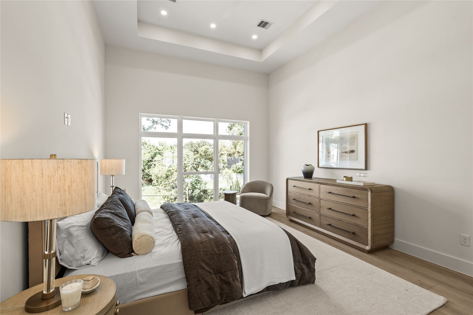 The primary bedroom in full. Tray ceiling with cove lighting, floor-to-ceiling glass, and a tree outside the window.