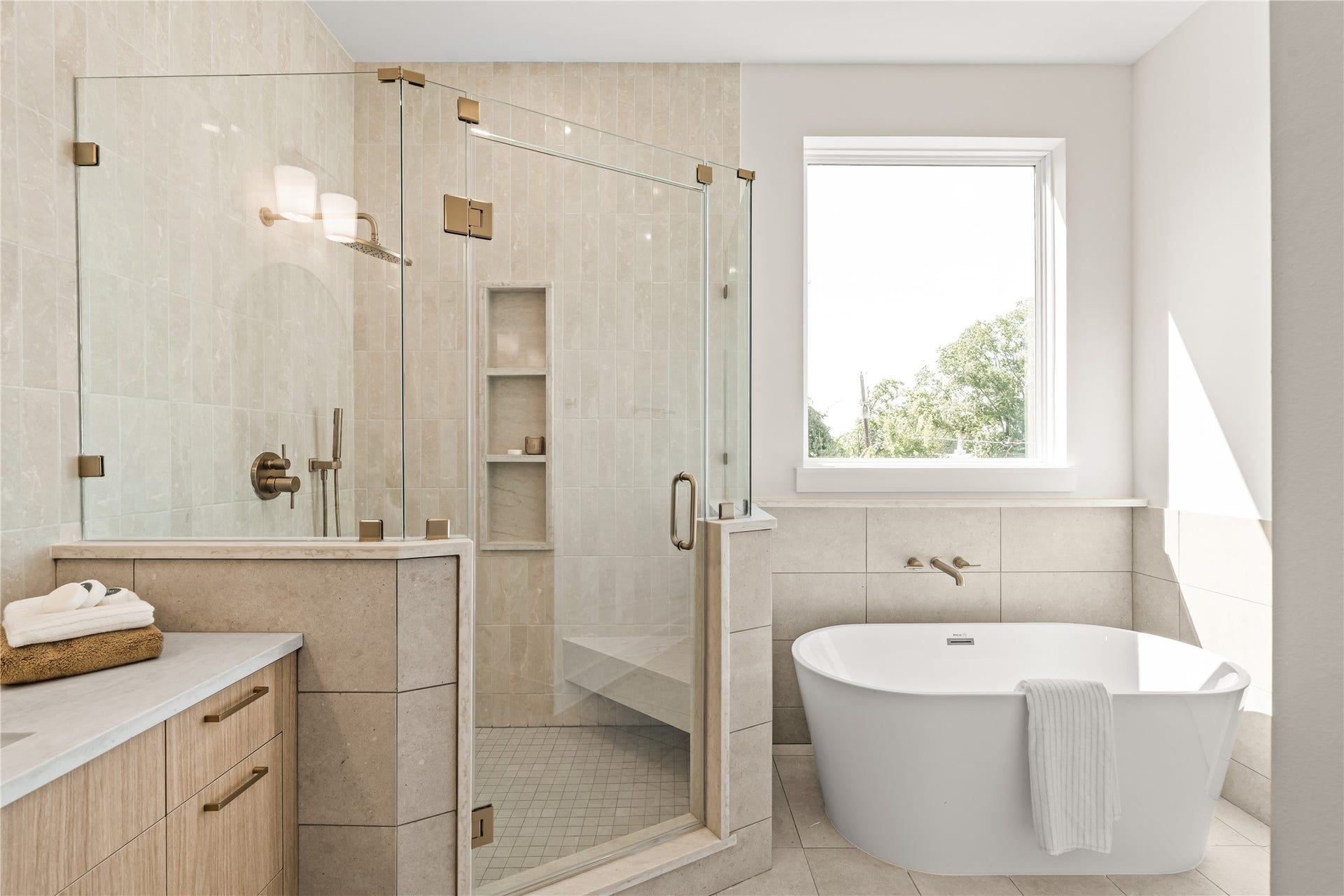 Freestanding soaking tub set under the window, frameless shower beside it. The primary bath in full, tile wrapping both.