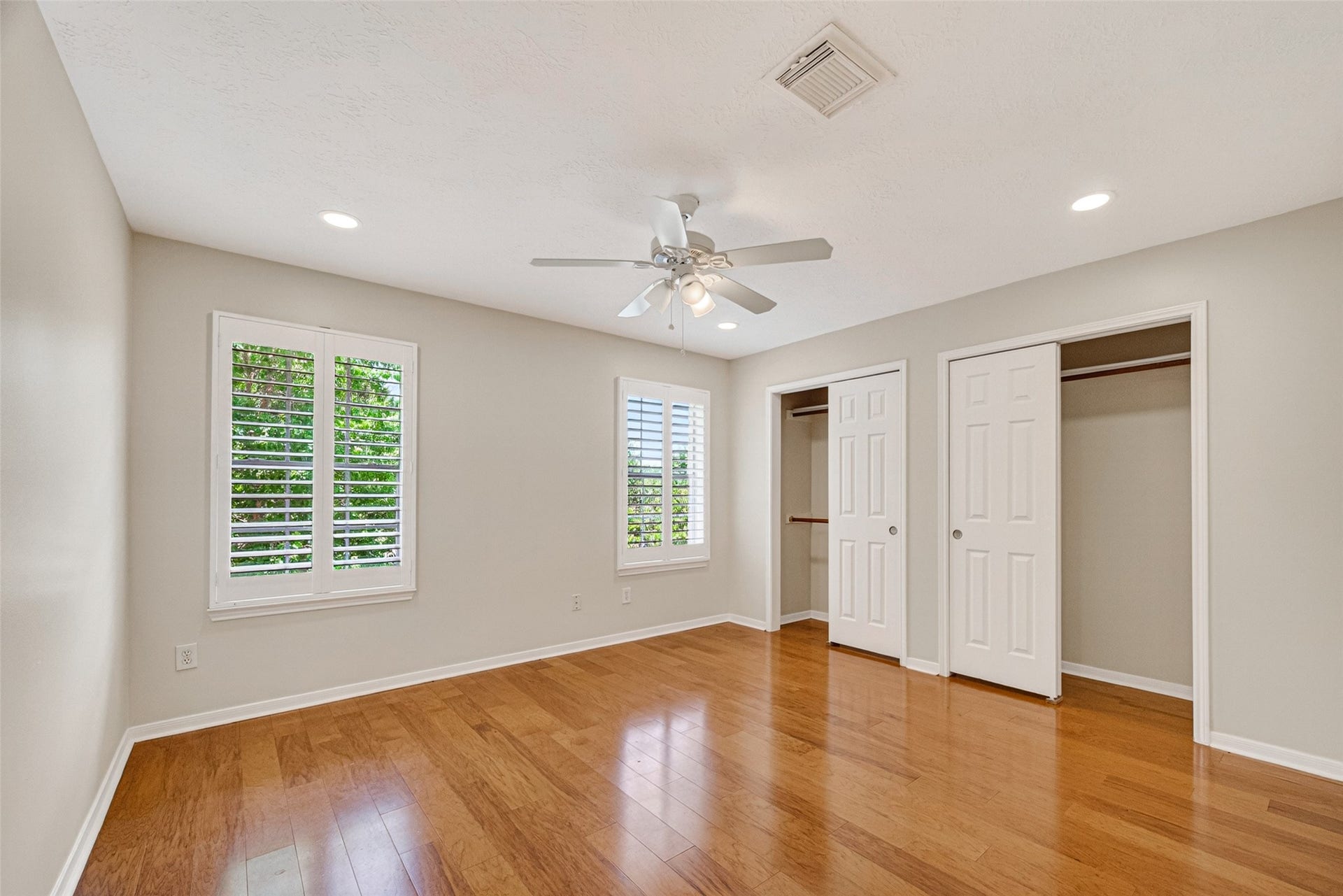 Bright and spacious bedroom featuring beautiful wood floors, large windows with plantation shutters, ample closet space, and a ceiling fan for comfort.