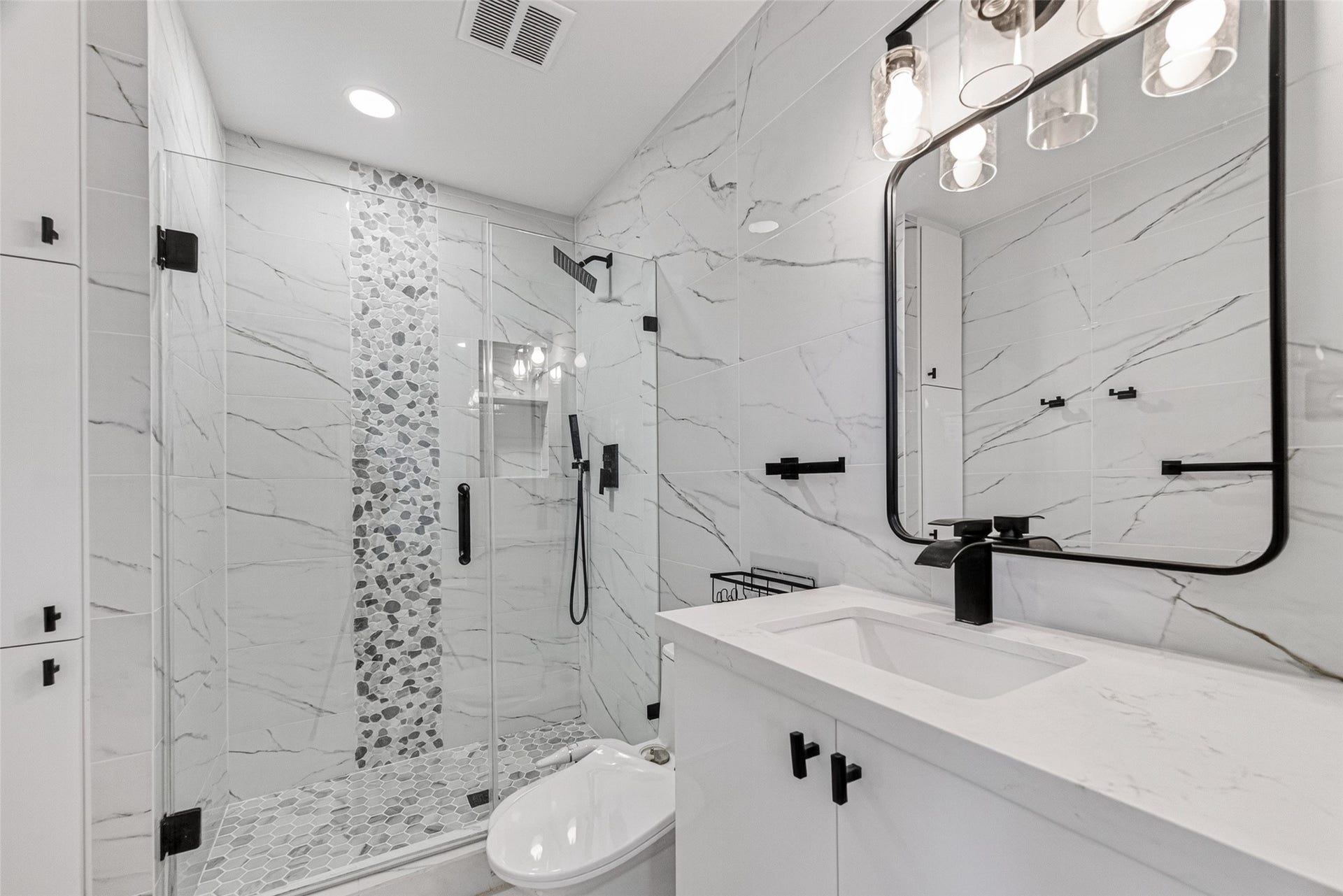 Bathroom # 1 (upstair) features a modern design with white marble-effect tiles, a sleek glass-enclosed shower with pebble accent strip, black fixtures, and a contemporary vanity with a large mirror and stylish lighting.