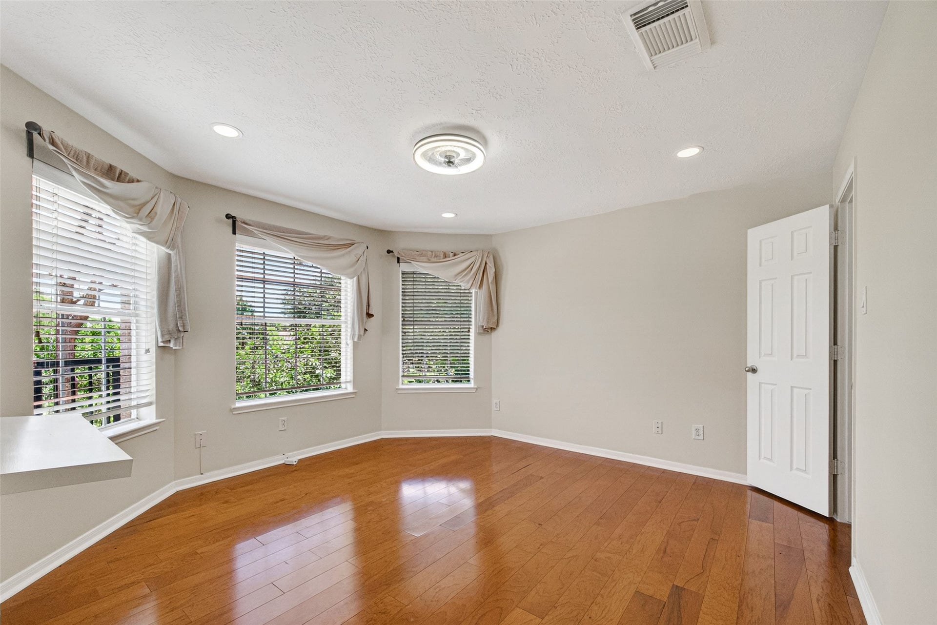 Bright and inviting bedroom featuring elegant wood floors, large windows with draped curtains, and ample natural light.