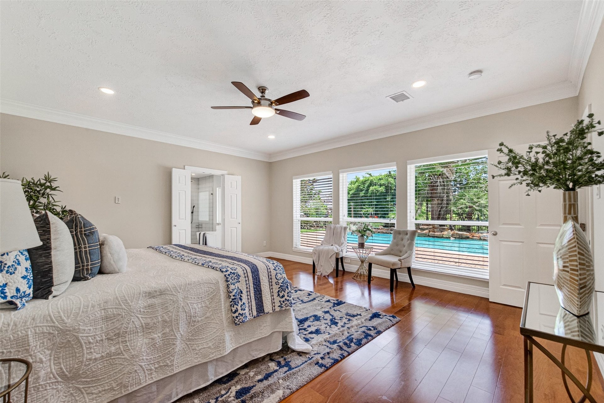 Primary bedroom features wood floors, a ceiling fan, and large windows offering a view of the pool. It includes an en-suite bathroom and ample natural light.