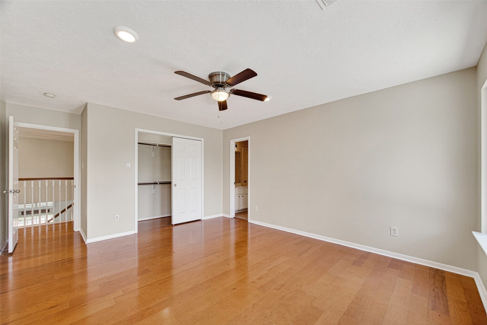 Spacious bedroom with wood floors, neutral walls, and a ceiling fan, featuring a large window for natural light and a walk-in closet.
