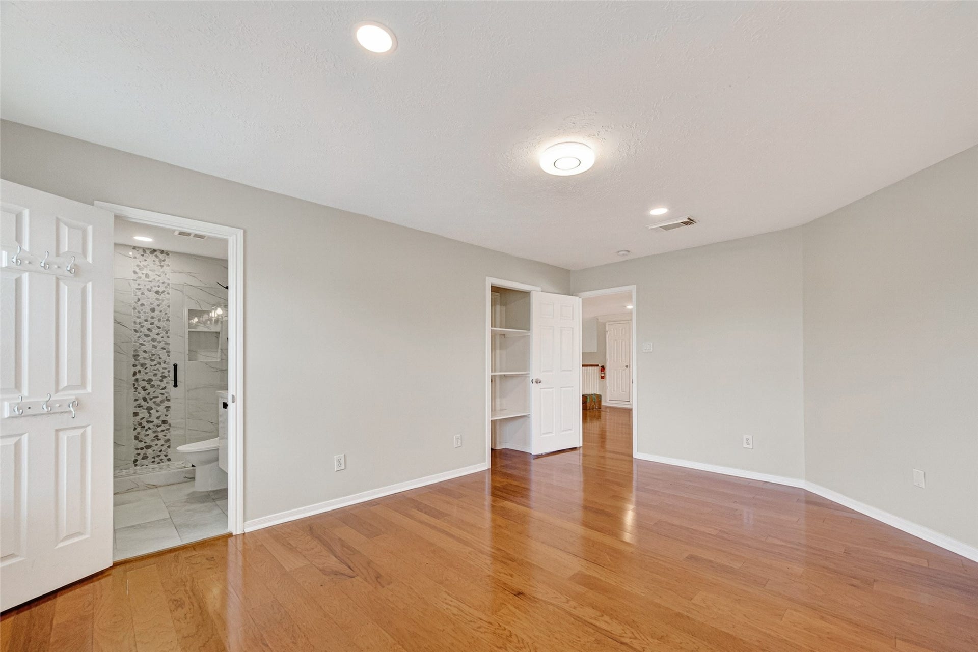 Spacious bedroom with polished wood floors and ample natural light, featuring an en-suite bathroom with modern tile accents and convenient built-in shelving.