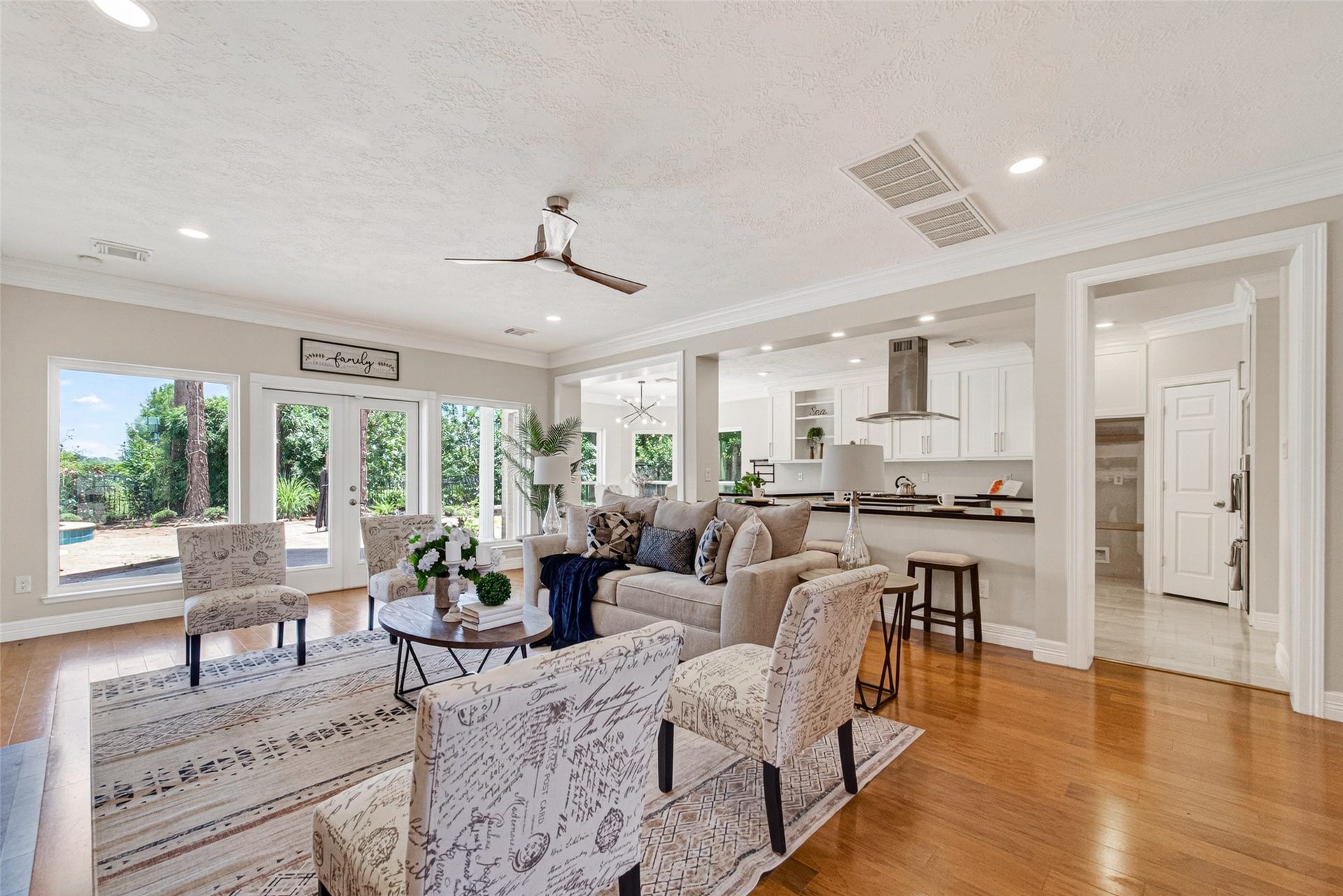 This spacious living area features an open floor plan with wood floors, modern ceiling fan, and ample natural light from large windows and French doors leading to a patio. The room seamlessly flows into a contemporary kitchen with white cabinetry and a breakfast bar, creating an inviting space for entertaining.
