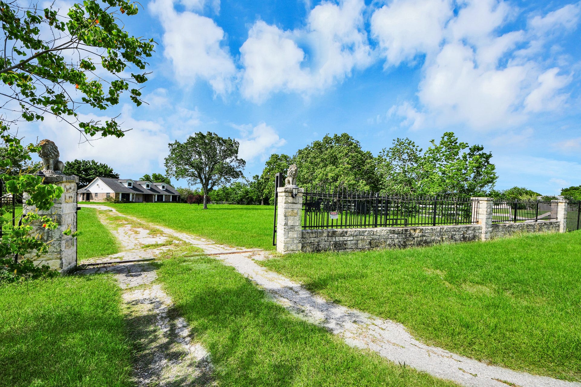 12746 Roy Road, Pearland, TX 77581 This photo features a charming country home set on a spacious green lawn. A long driveway leads to the house, passing through a stone gate with decorative statues. The property is surrounded by mature trees and open sky, offering a sense of privacy and tranquility.