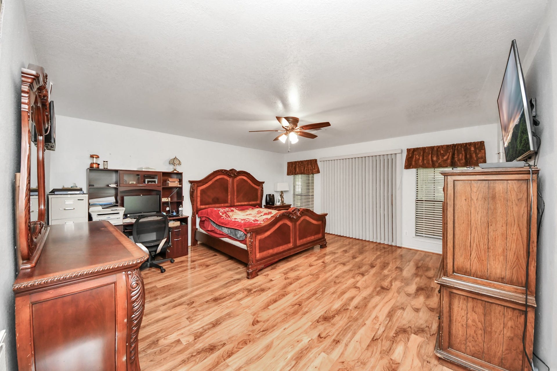 12746 Roy Road, Pearland, TX 77581 This spacious bedroom features elegant hardwood flooring, a large wooden bed, and matching furniture. It includes a home office setup with a desk and chair, a ceiling fan, and ample natural light from sliding glass doors and windows with blinds.