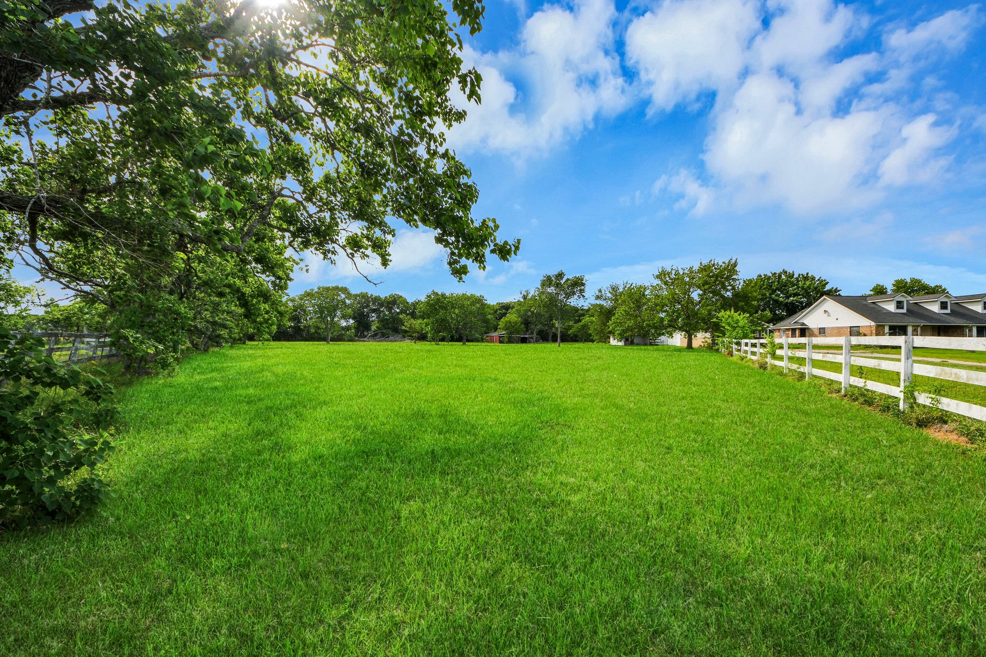 12746 Roy Road, Pearland, TX 77581 This photo showcases a spacious, lush green field bordered by white fencing and trees, under a clear blue sky, offering a serene and private setting ideal for building a dream home.