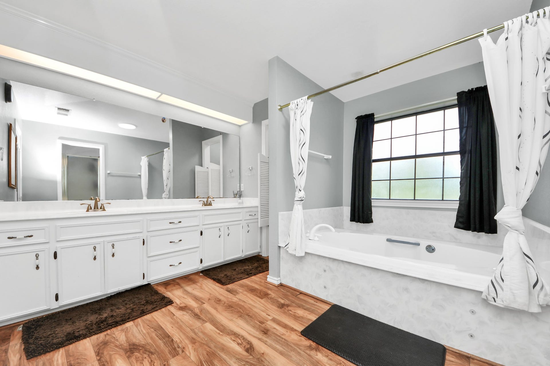 12746 Roy Road, Pearland, TX 77581 This spacious bathroom features a large vanity with dual sinks, ample cabinet storage, and elegant fixtures. A luxurious soaking tub is set beneath a wide window, providing natural light and a relaxing atmosphere. The room is enhanced by wood-look flooring and soft, neutral wall colors.