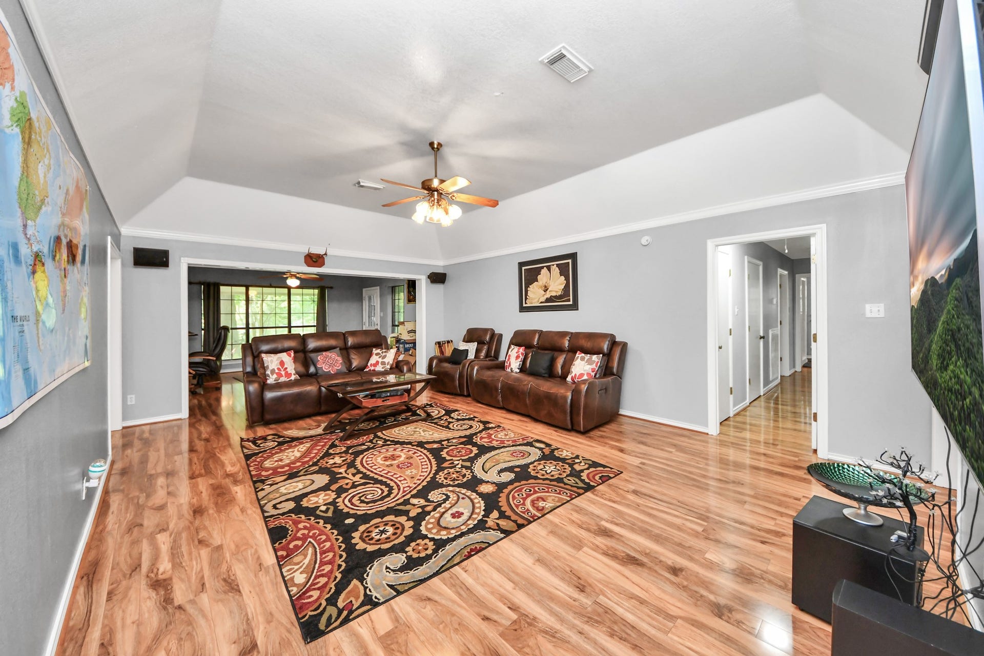 12746 Roy Road, Pearland, TX 77581 This spacious living room features polished wood flooring, a ceiling fan, and ample natural light from large windows. It includes a cozy seating area with leather couches and a colorful area rug. The room has a modern, open feel with neutral walls and direct access to a hallway and adjacent dining area.