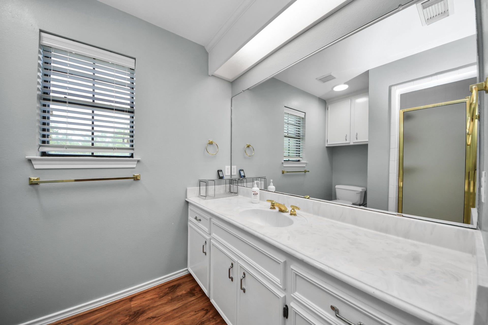 12746 Roy Road, Pearland, TX 77581 This bathroom features a large marble-topped vanity with dual sinks, a spacious mirror, and gold fixtures. It has a glass-enclosed shower, white cabinetry, and a window with blinds, offering a clean and modern look.