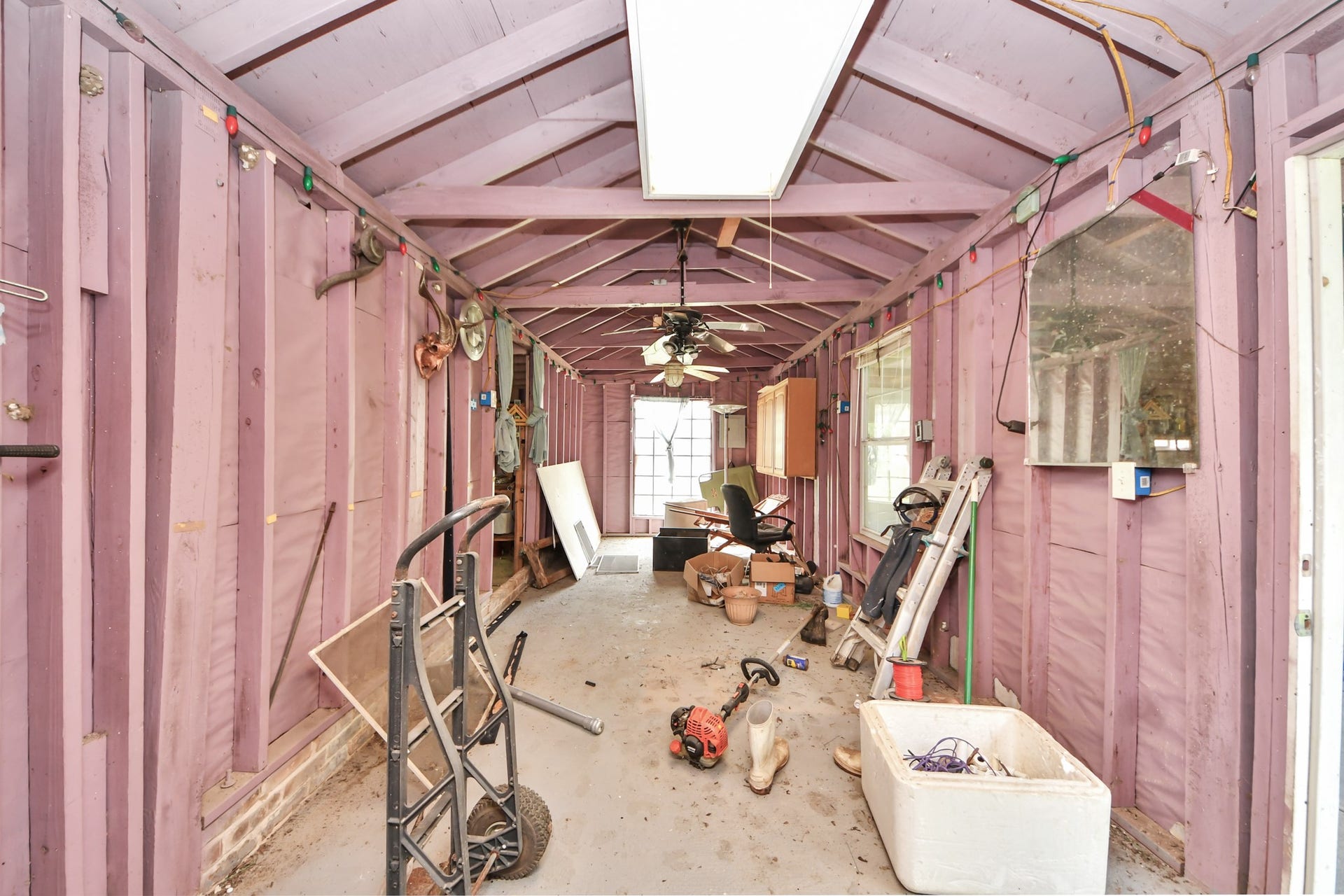 12746 Roy Road, Pearland, TX 77581 This photo shows a long, narrow utility or storage room with exposed beams and walls painted in a light purple. The space contains various tools and equipment, with a ceiling fan and fluorescent lighting. Natural light enters through a window at the far end.