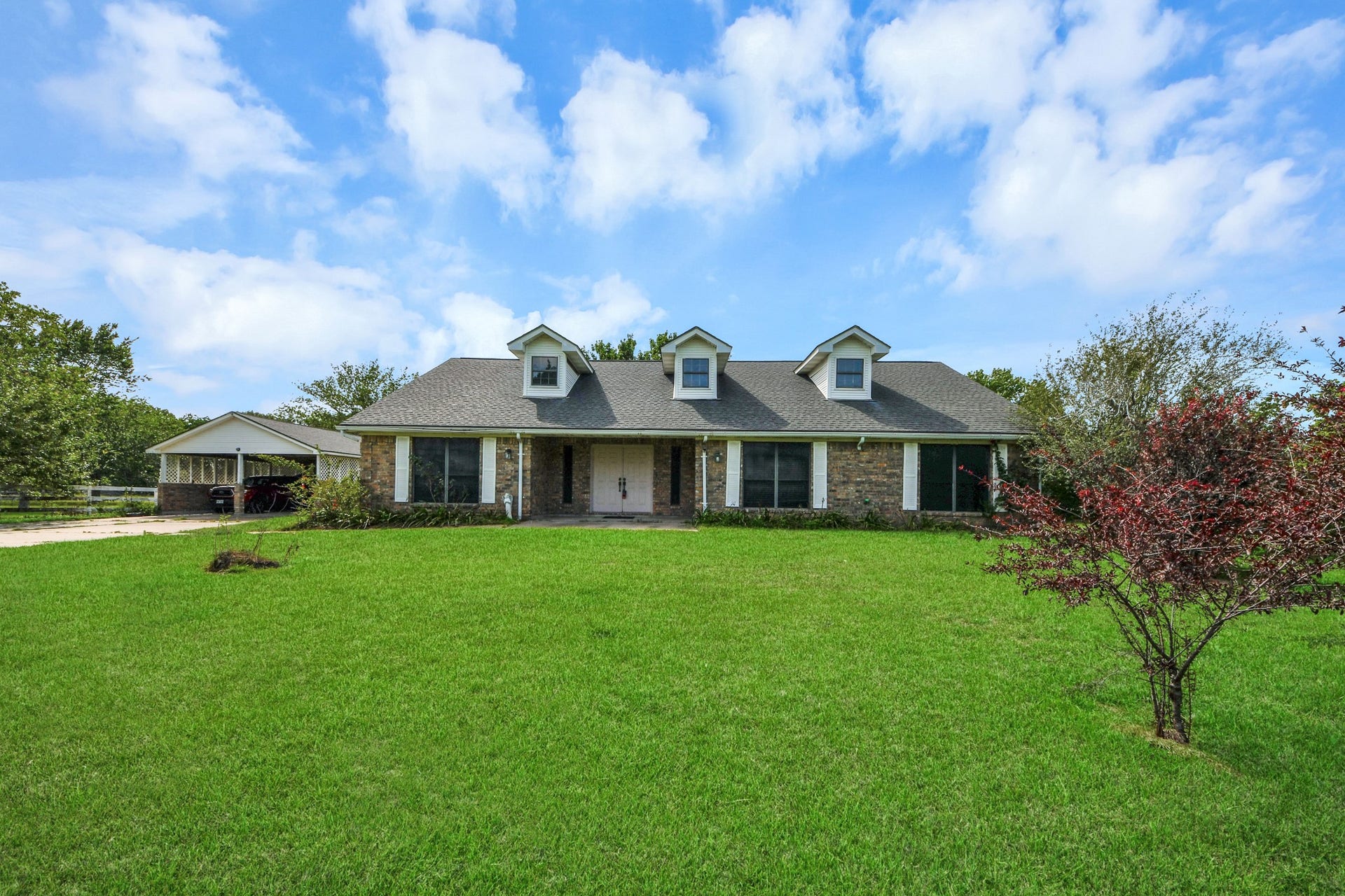 12746 Roy Road, Pearland, TX 77581 Charming single-story brick home with a large front lawn, featuring three dormer windows on the roof. It includes a detached garage and is surrounded by greenery, offering a peaceful suburban feel.