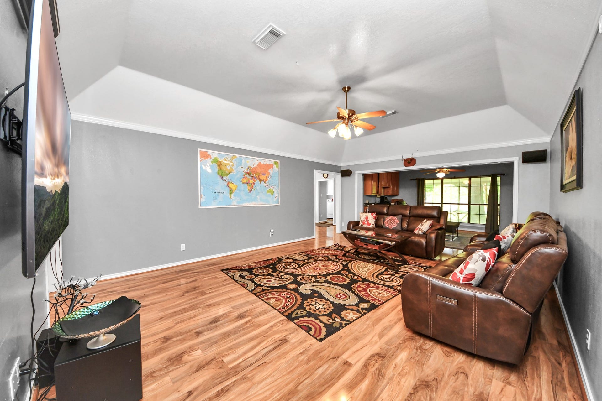 12746 Roy Road, Pearland, TX 77581 Spacious living room with vaulted ceilings, featuring hardwood floors and a large ceiling fan. The room is equipped with comfortable leather seating, a wall-mounted TV, and a colorful area rug. A map adorns the wall, and it opens into a bright kitchen/dining area with ample natural light.