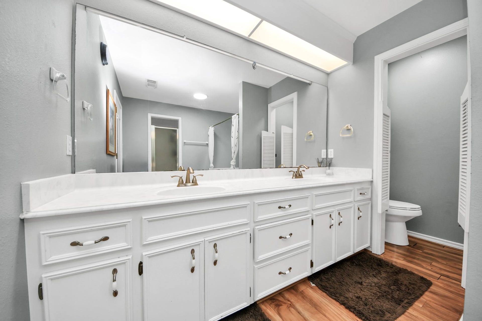 12746 Roy Road, Pearland, TX 77581 This bathroom features a spacious dual-sink vanity with white cabinetry and brass fixtures, a large mirror, wood flooring, and a separate toilet area for added privacy.