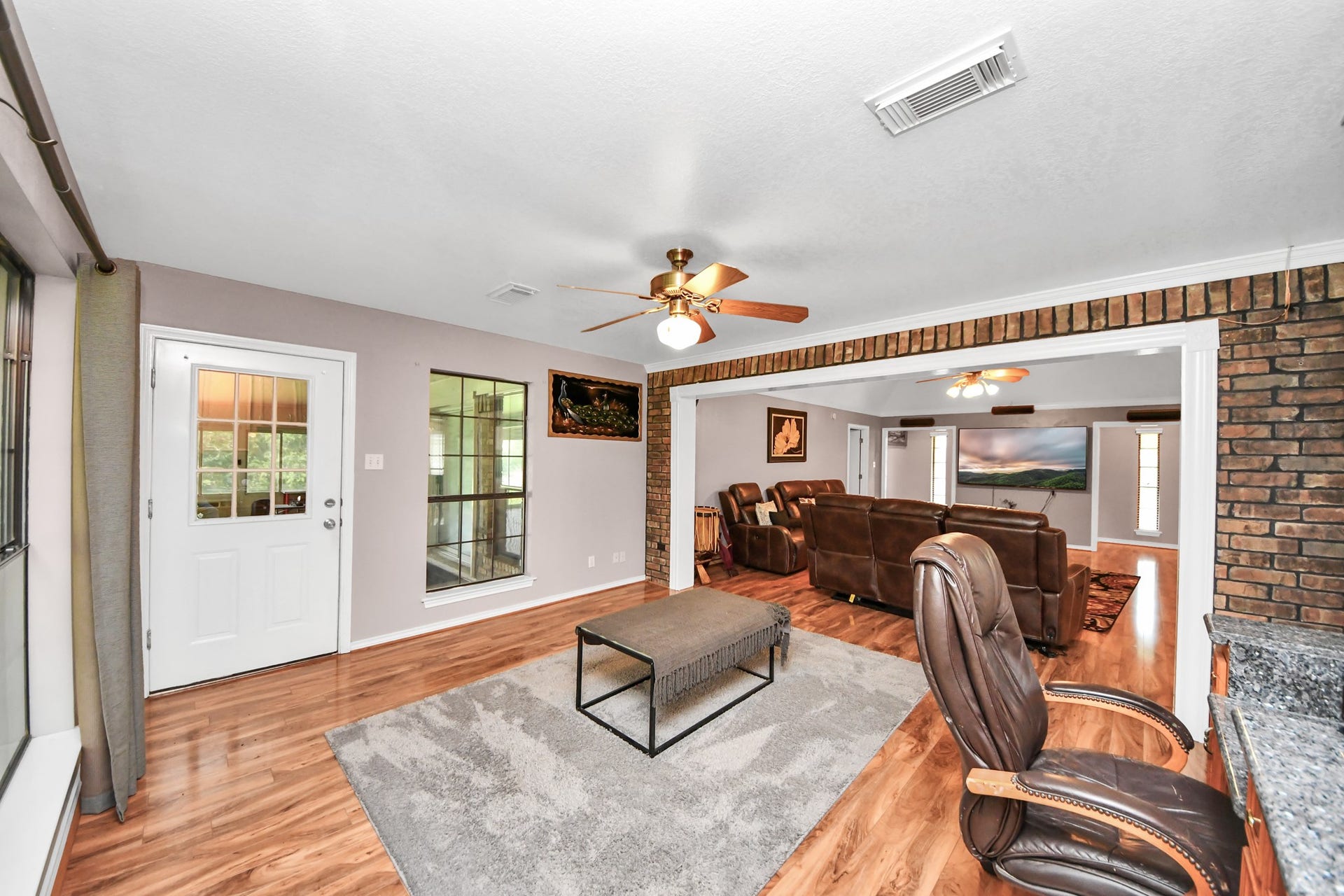 12746 Roy Road, Pearland, TX 77581 This spacious living area features warm wood flooring, a cozy brick accent wall, and abundant natural light. It includes a ceiling fan for comfort, an inviting open layout with a comfortable seating area, and a view of the outdoors through large windows.