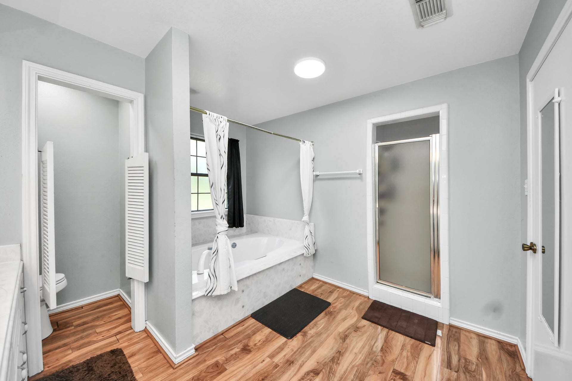 12746 Roy Road, Pearland, TX 77581 This spacious bathroom features a large soaking tub with a curtain, a separate shower with frosted glass, and elegant wood flooring. Natural light pours in through a window, creating a bright and inviting atmosphere.