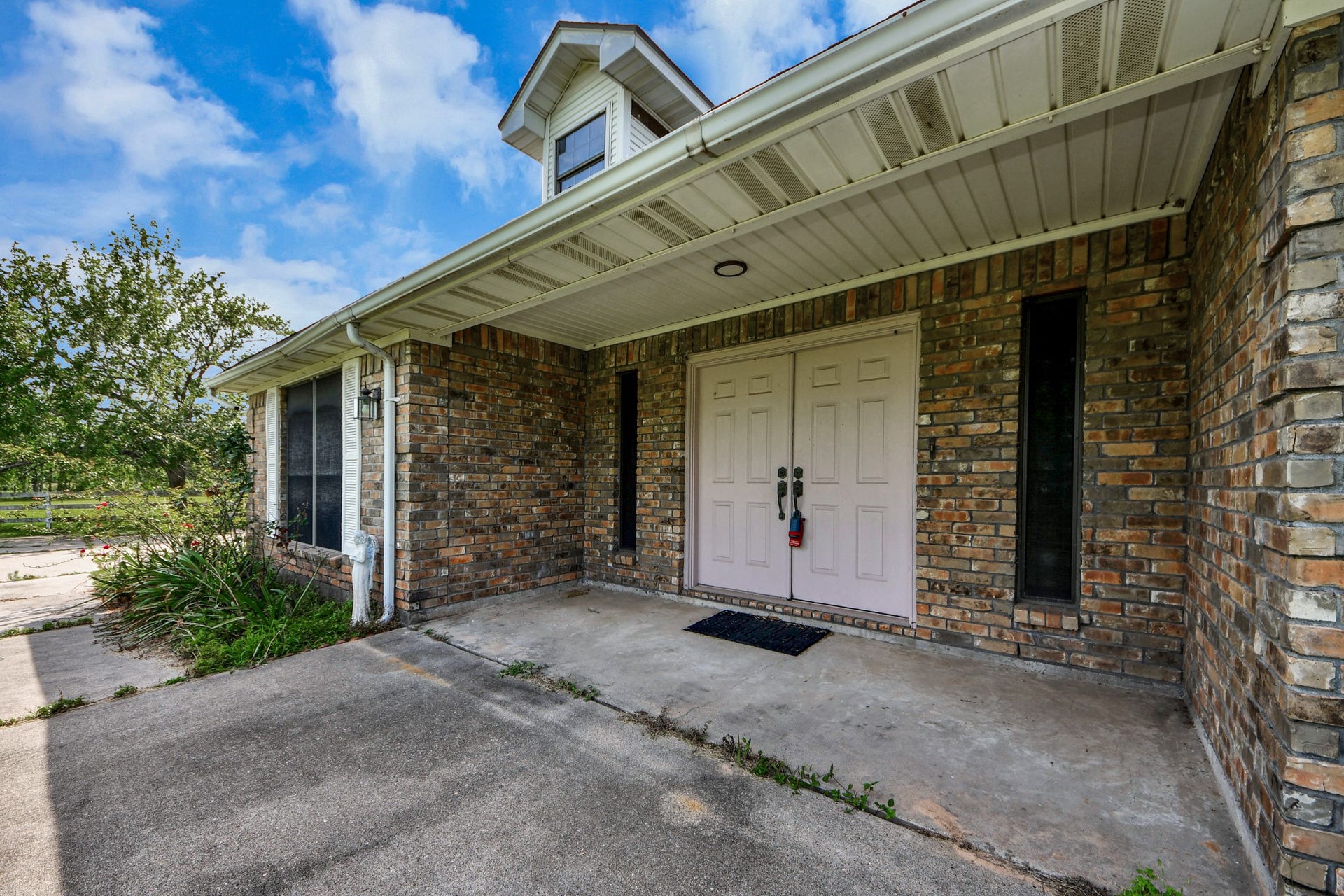 12746 Roy Road, Pearland, TX 77581 This photo showcases the front entrance of a home featuring a covered porch and double doors. The exterior is composed of brick, providing a classic and sturdy appearance. The walkway is bordered by some greenery and mature trees, offering a welcoming approach to the house.