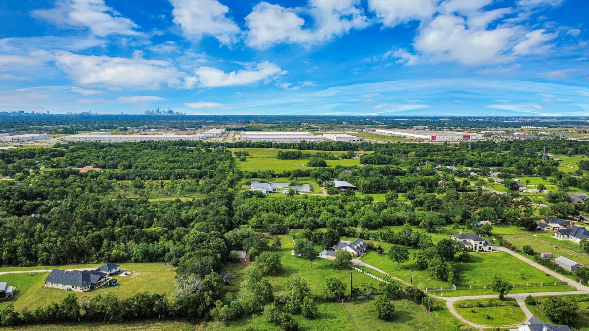 12746 Roy Road, Pearland, TX 77581 This aerial photo showcases a spacious, green area with scattered homes, offering a peaceful rural setting. The landscape is lush with trees and open fields, and a distant view of a city skyline is visible, suggesting convenient access to urban amenities.