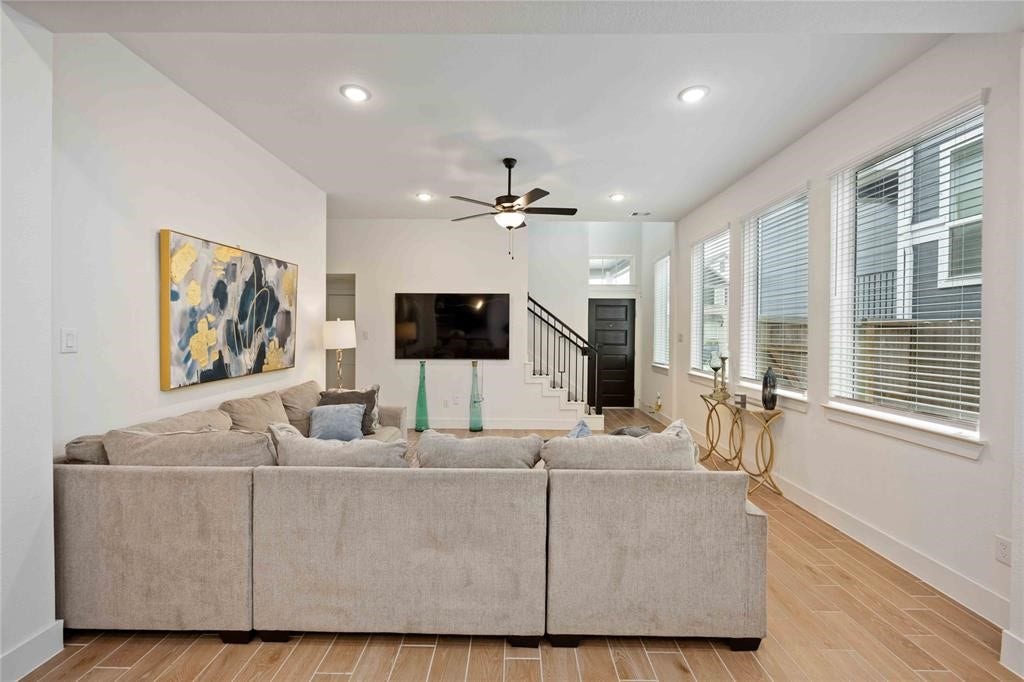Virtually Staged - Bright and inviting living room featuring a large sectional sofa, wall-mounted TV, and ceiling fan. Natural light pours in through multiple windows, highlighting the modern decor and open floor plan.