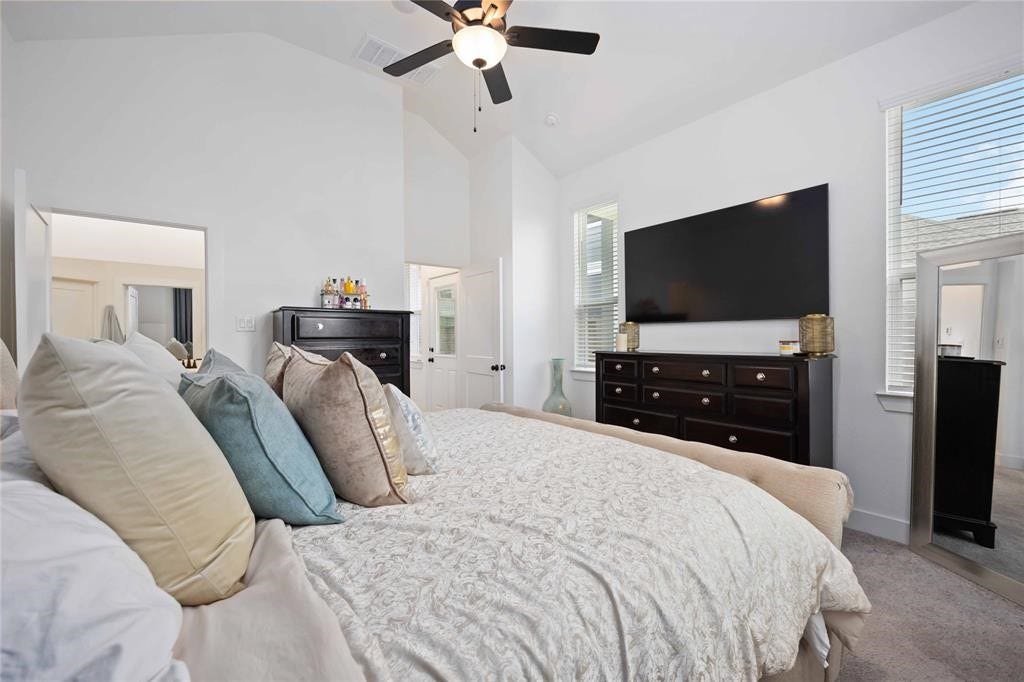 Virtually Staged - This is a bright, spacious bedroom featuring a large bed with plush pillows, a ceiling fan, and a wall-mounted TV. The room has ample natural light from two windows, a dark wood dresser, and a stylish floor mirror, creating a cozy and inviting atmosphere.