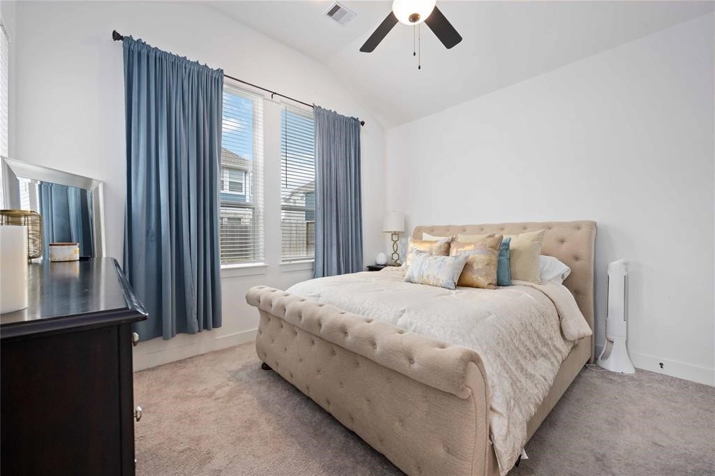 Virtually Staged - Cozy bedroom with a plush tufted bed, neutral carpeting, and soft blue curtains. The room features a ceiling fan and ample natural light from double windows, creating a serene and inviting atmosphere.