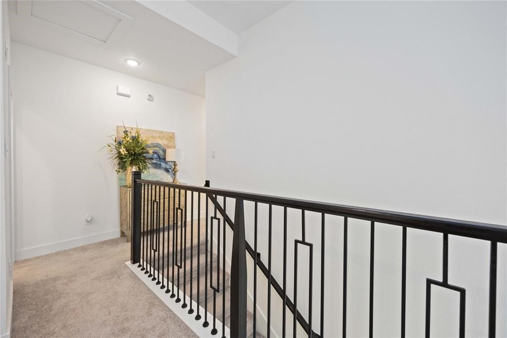 Virtually Staged - This photo shows a bright hallway with plush carpeting, a modern black metal railing, and a decorative plant on a console table against a white wall.