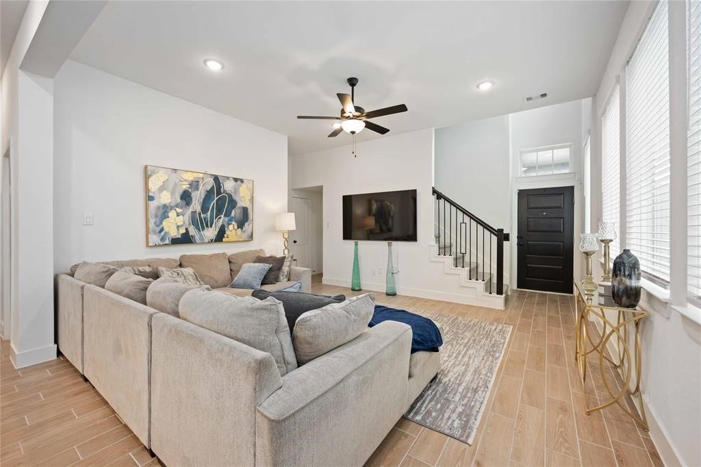 Virtually Staged - Spacious living room with modern decor, featuring a large sectional sofa, wall-mounted TV, and stylish ceiling fan. The room has light wood flooring, abundant natural light from large windows, and a staircase leading to the upper level.
