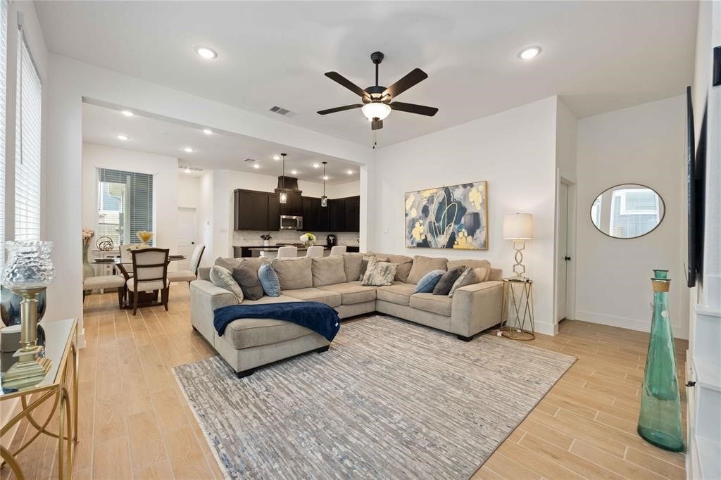 Virtually Staged - This inviting open-concept living area features a spacious sectional sofa, modern decor, and a ceiling fan. The room seamlessly connects to a dining area and a sleek kitchen with dark cabinetry and stainless steel appliances, all under warm recessed lighting.
