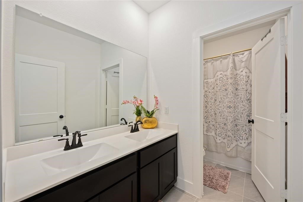 Virtually Staged - This bathroom features a modern double vanity with dark cabinetry and sleek fixtures, a large mirror, and a shower with a patterned curtain. The space is bright and inviting with white walls and tile flooring.