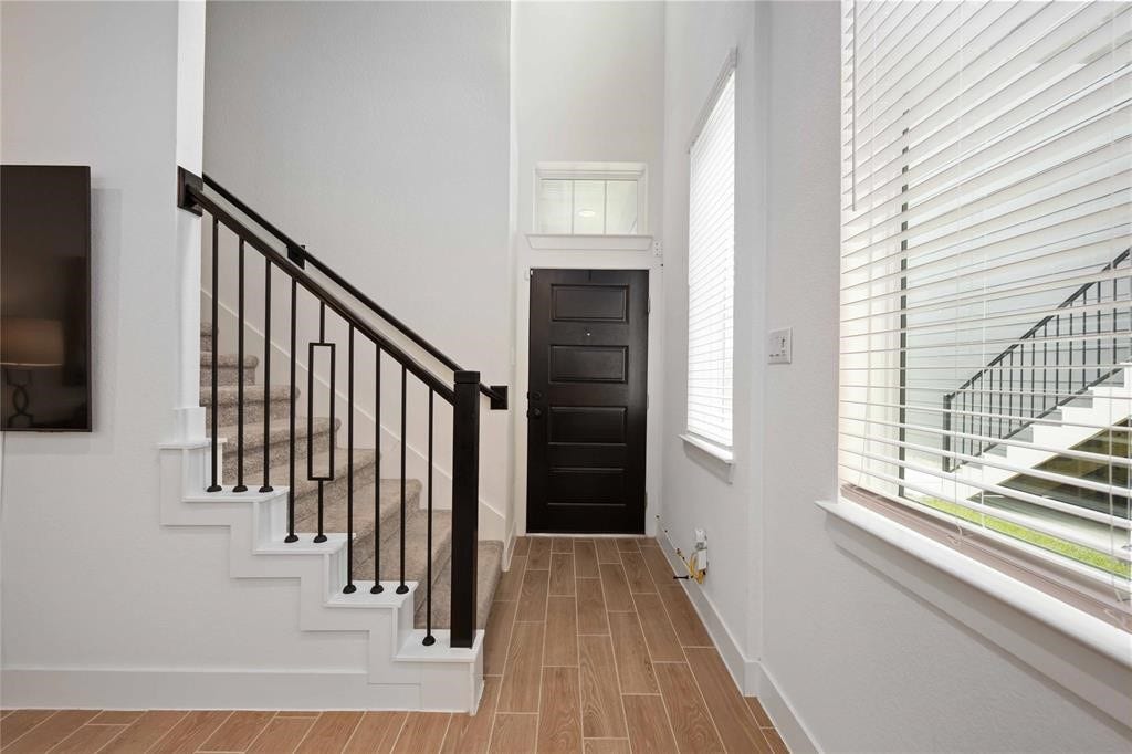 Virtually Staged - Bright entryway with high ceilings, featuring a modern staircase with black railings. The space includes large windows with blinds, and a sleek black front door. Wood-look tile flooring adds warmth and style.