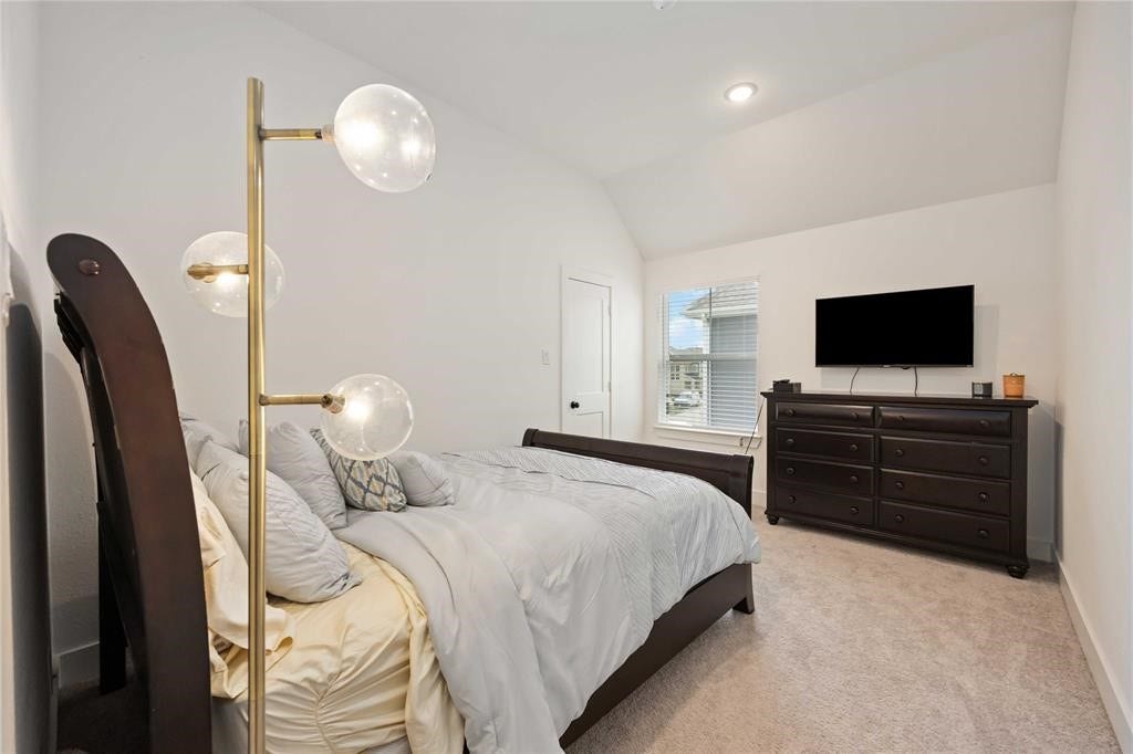 Virtually Staged - This cozy bedroom features a vaulted ceiling, soft carpeting, and a large window for natural light. It includes a modern floor lamp, a comfortable bed with plush bedding, and a dark wood dresser topped with a mounted TV, creating a relaxing ambiance.