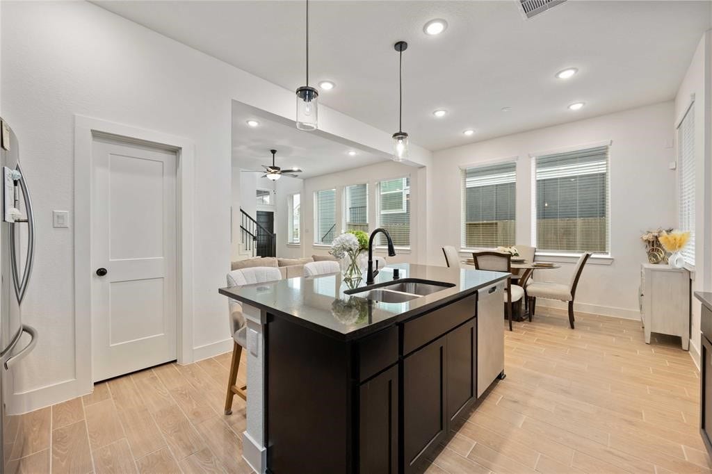 Virtually Staged - This photo showcases a modern, open-concept kitchen and dining area with sleek lighting. The kitchen features a dark island with a sink, complemented by light wood flooring. Large windows provide ample natural light, and there's a cozy living space visible in the background.