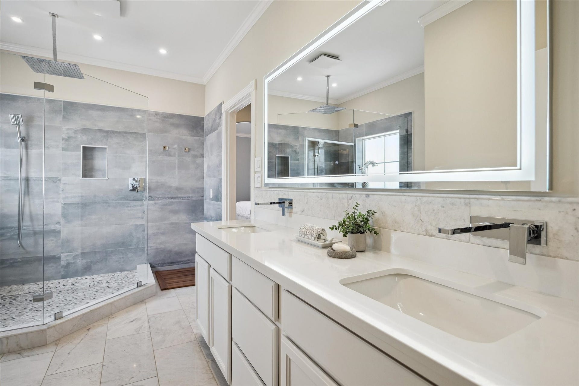 5717 Val Verde Street, #C, Houston, TX 77057 This modern bathroom features a spacious glass-enclosed shower with sleek gray tiles, dual rainfall showerheads, and a pebble stone floor. The double vanity has a marble countertop, contemporary fixtures, and an illuminated mirror, offering a luxurious and functional space.