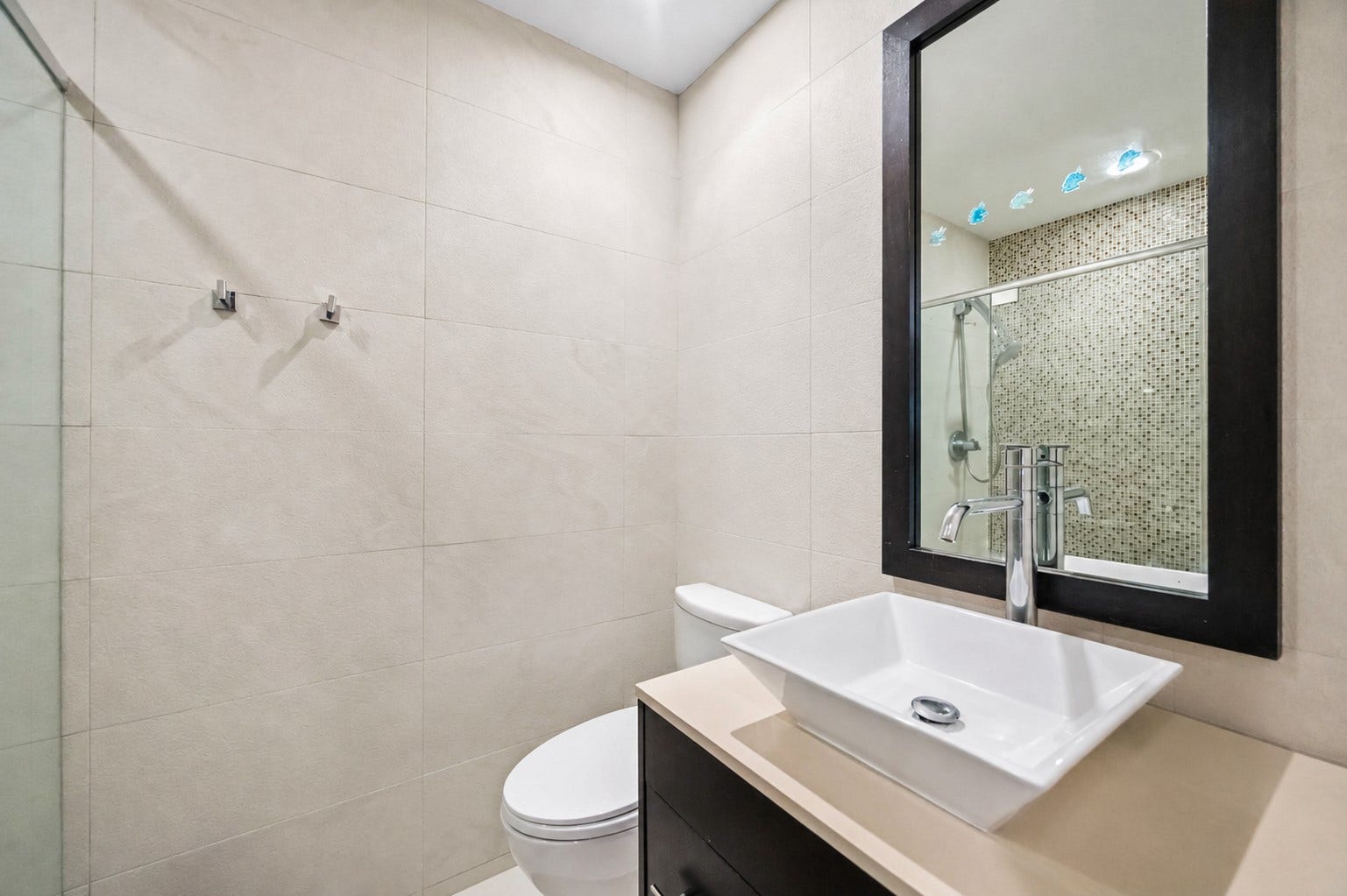 7610 Guinevere Drive, Sugar Land, TX 77479 This bathroom spaBathroom with neutral tile, dark floating vanity, white countertop, vessel sink, and framed mirror.ce features premium neutral wall tiling, a dark floating vanity cabinet with a sleek white countertop, and a vessel sink beneath a dark-framed mirror.