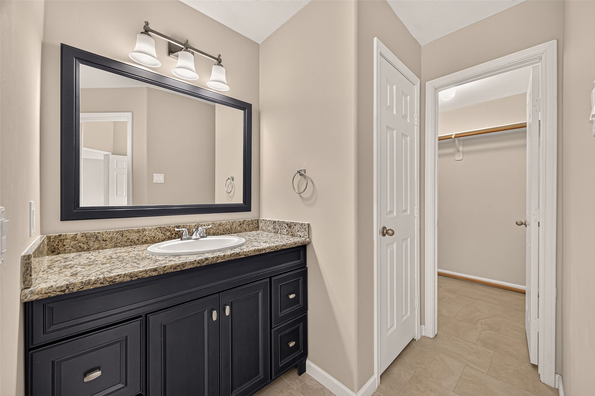 Sophisticated bathroom with a sleek black vanity and granite countertop, enhanced by a large framed mirror and modern lighting, next to a spacious walk-in closet for ample storage.