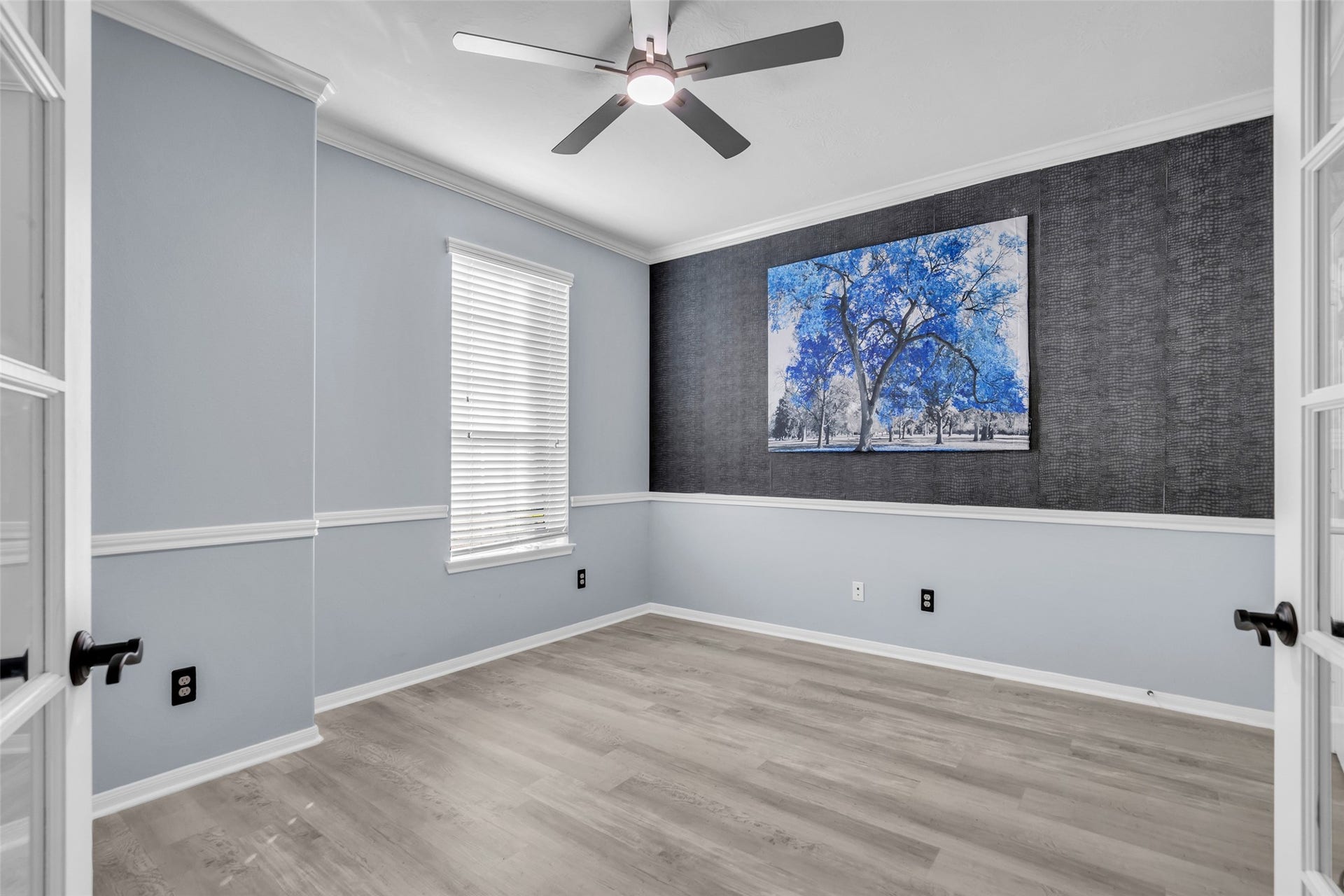 8121 Tranquil Lake Way, Conroe, TX 77385 Bright and inviting office featuring elegant gray walls with white trim, stylish wood flooring, and a contemporary ceiling fan. A striking piece of art adds a pop of color, perfect for a home office or cozy retreat.