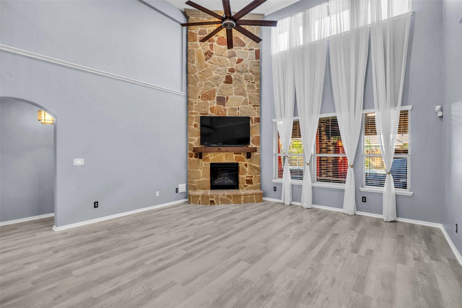 8121 Tranquil Lake Way, Conroe, TX 77385 Bright and spacious living room featuring soaring ceilings, a stunning stone fireplace, and large windows adorned with elegant sheer curtains, creating a warm and inviting atmosphere.