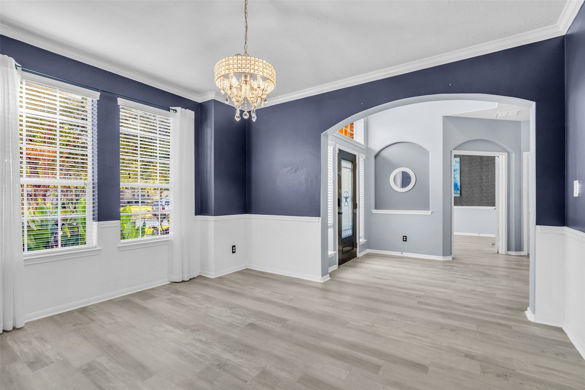 8121 Tranquil Lake Way, Conroe, TX 77385 Elegant formal dining area featuring bold navy walls, white wainscoting, and a sparkling chandelier, complemented by abundant natural light from large windows and stylish archways leading to adjacent rooms.