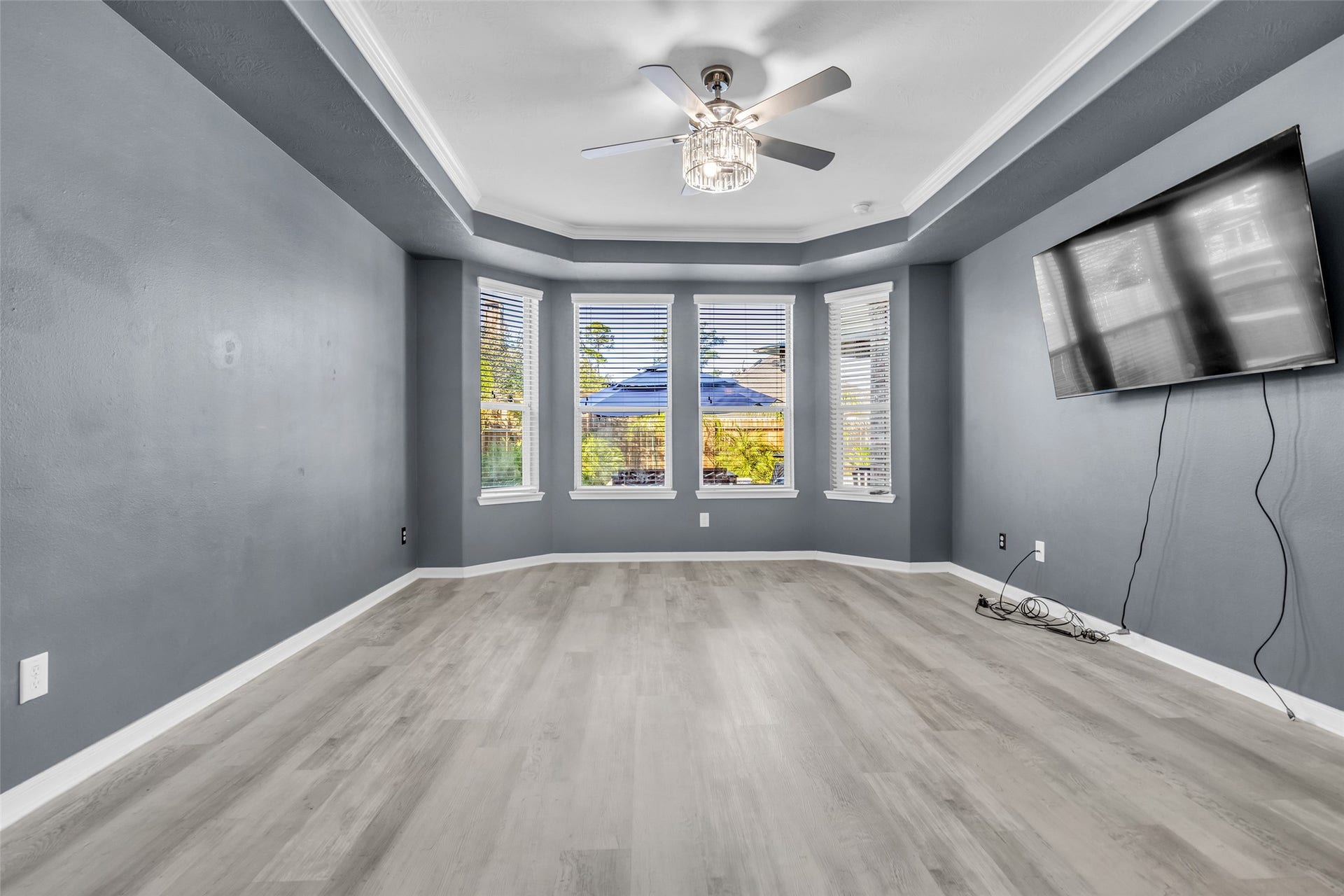 8121 Tranquil Lake Way, Conroe, TX 77385 Spacious primary bedroom featuring elegant tray ceiling, chic gray walls, and abundant natural light from large bay windows, complemented by modern flooring and a ceiling fan.