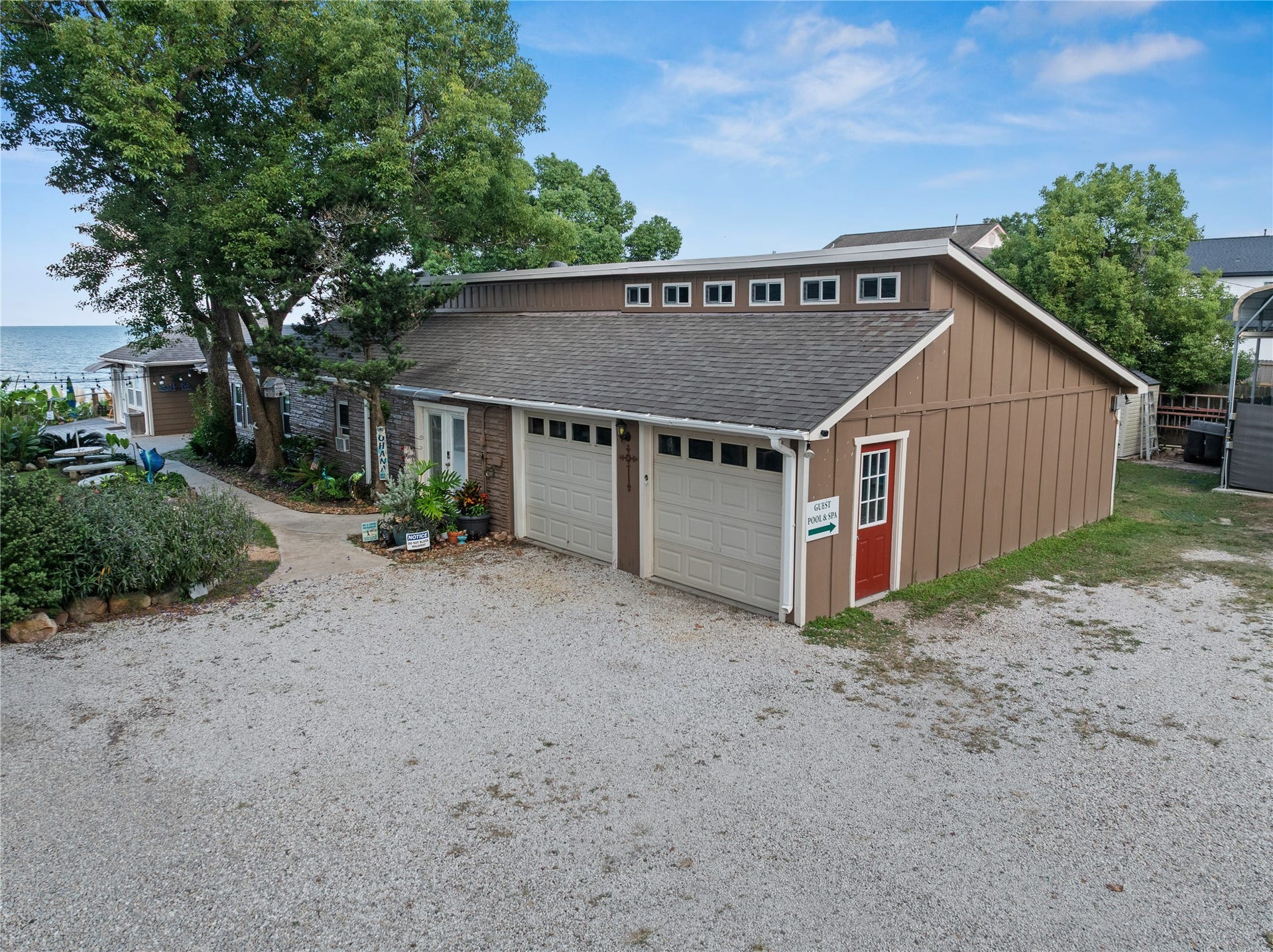 3709 Bayshore Drive, #1-12, Bacliff, TX 77518 This photo showcases the charming waterfront Ohana House with a rustic exterior, featuring a two-car garage and mature trees. A gravel driveway leads to the property, which offers scenic water views and a cozy atmosphere. Perfect for those seeking a peaceful retreat.