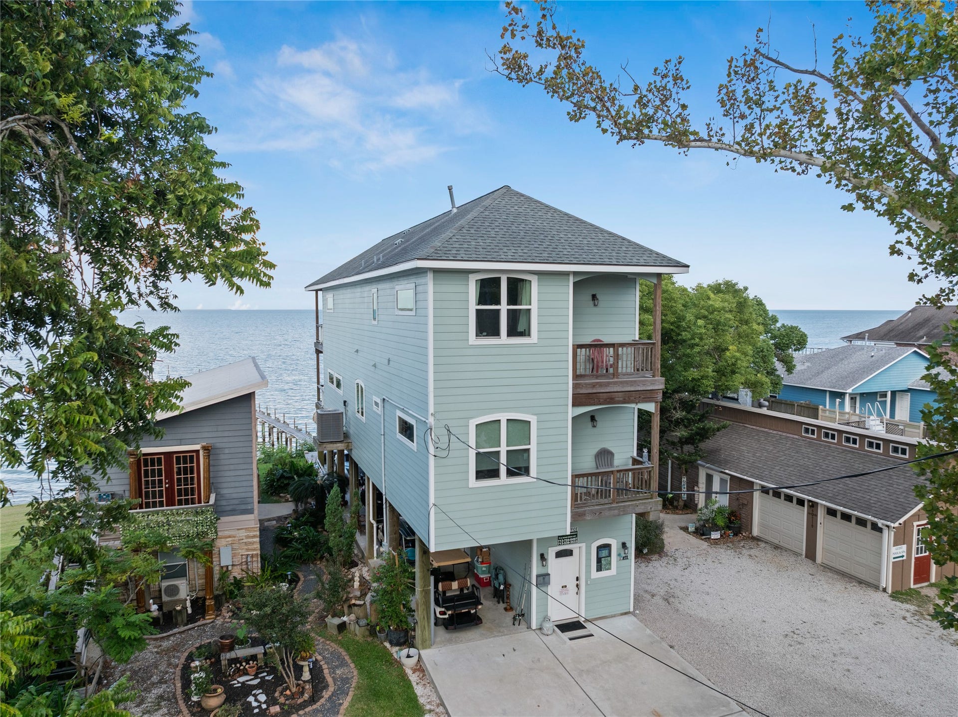 3709 Bayshore Drive, #1-12, Bacliff, TX 77518 This photo features the charming, elevated coastal home with two stories, featuring a light blue exterior and two balconies with waterfront views. It's nestled among trees with a small landscaped garden and offers bay side living with direct access to the waterfront.