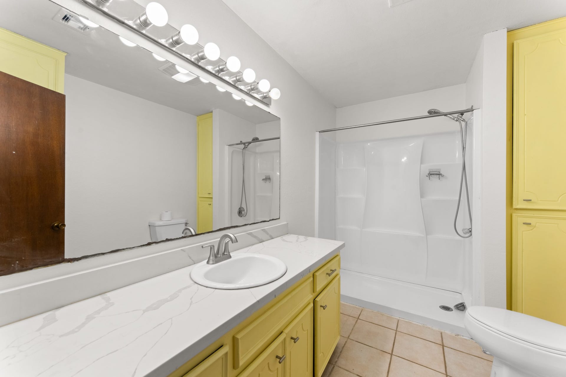 11325 32nd Street, Santa Fe, TX 77510 Spacious bathroom featuring a long vanity with marble-look countertops, a large wall mirror, cheerful yellow cabinetry, additional built-in storage, and a walk-in shower with dual showerheads.