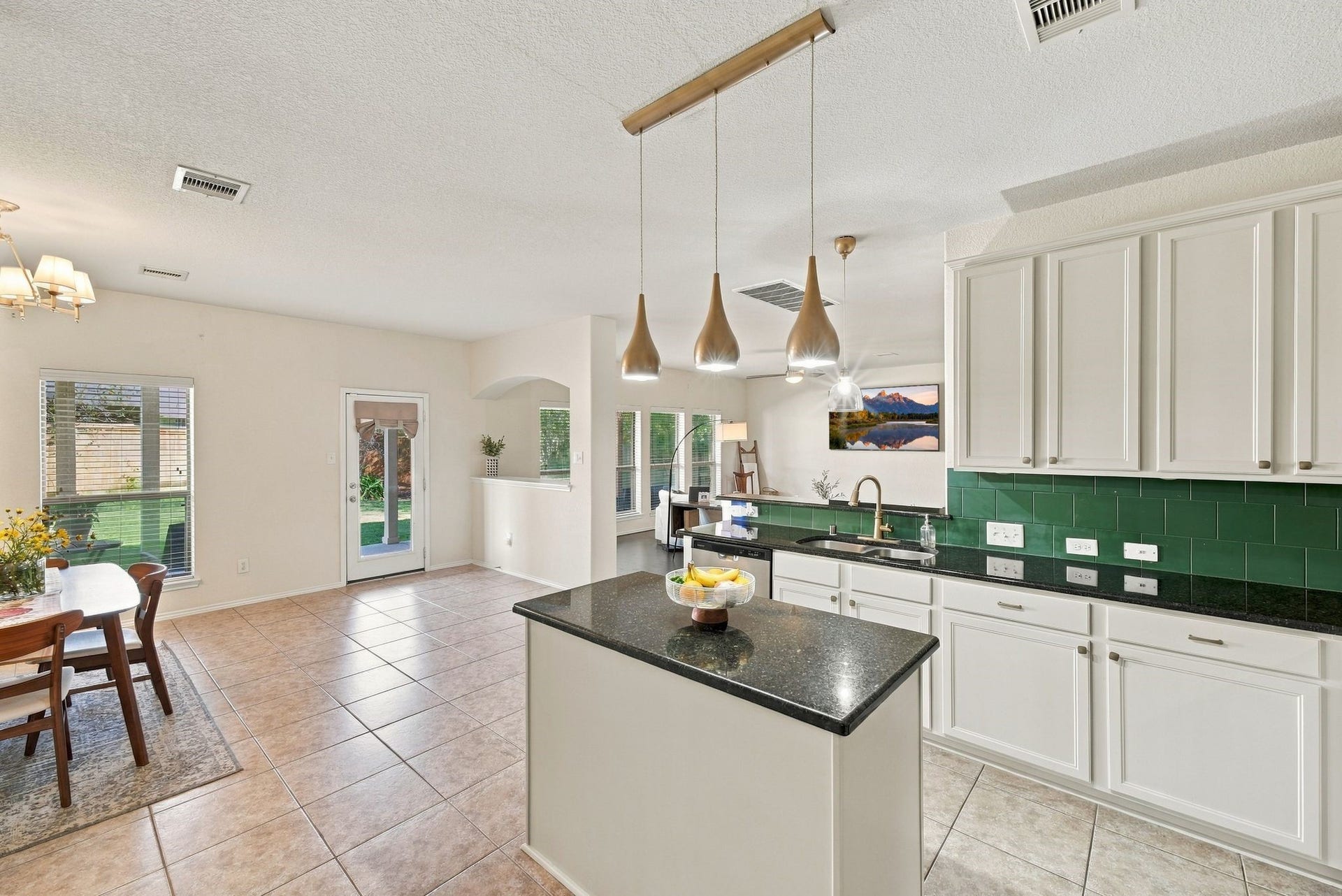 26037 Kings Mill Crest Drive, Kingwood, TX 77339 Spacious kitchen featuring sleek white cabinetry, a dark granite countertop island with pendant lighting, and a vibrant green backsplash. The open layout flows seamlessly into a bright dining area with large windows and a view of the backyard.