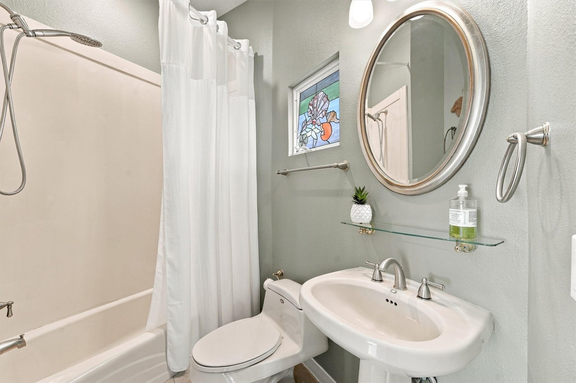4207 Barataria Court, Galveston, TX 77554 A colorful stained glass leans against the window adding seaside charm to this bath. A tub-and-shower combination, pedestal sink, and glass display shelf create a convenient place to freshen up.