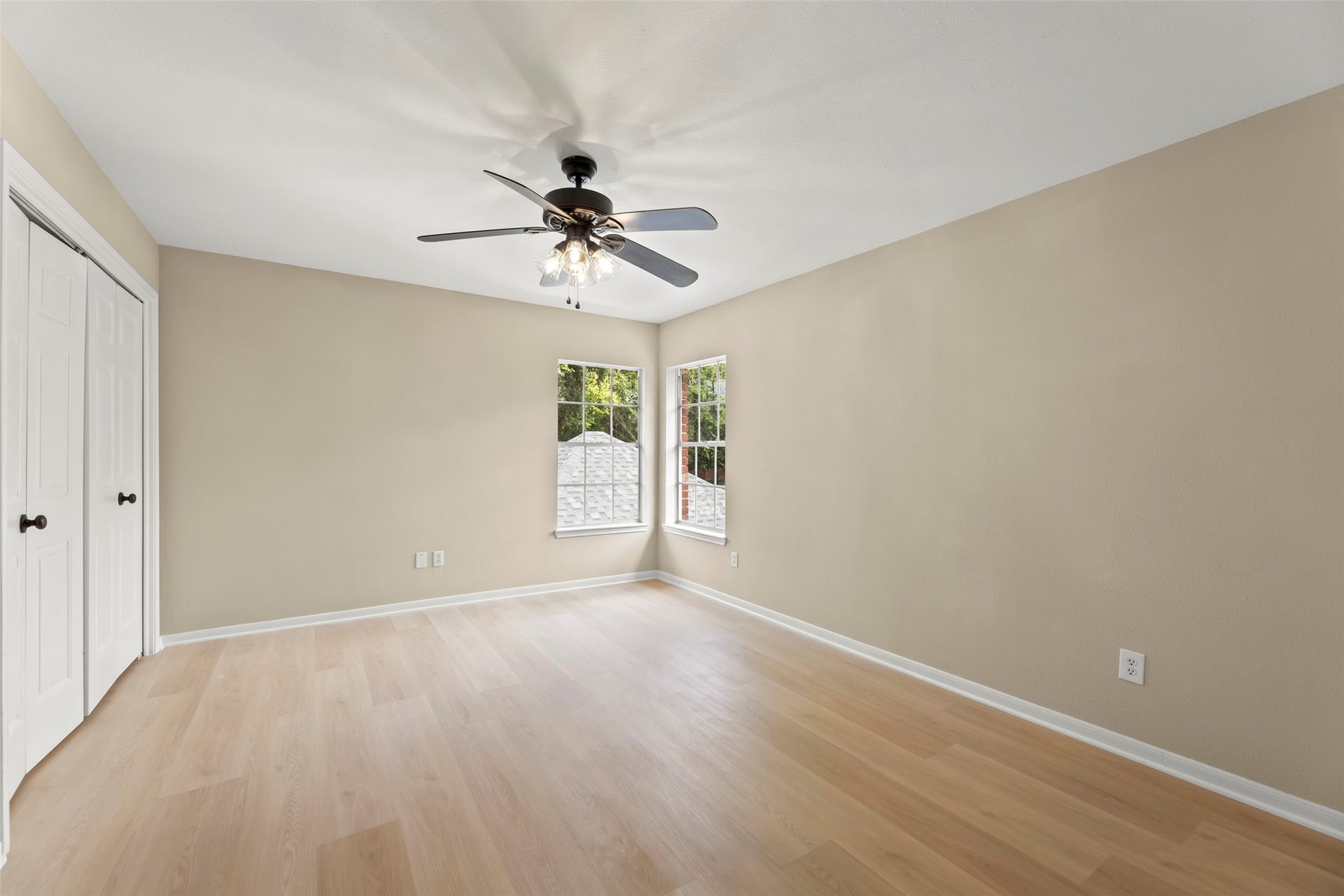 Vivid, spacious room with light wood flooring, neutral walls, and a ceiling fan. Features two windows for natural light and a closet with double doors.