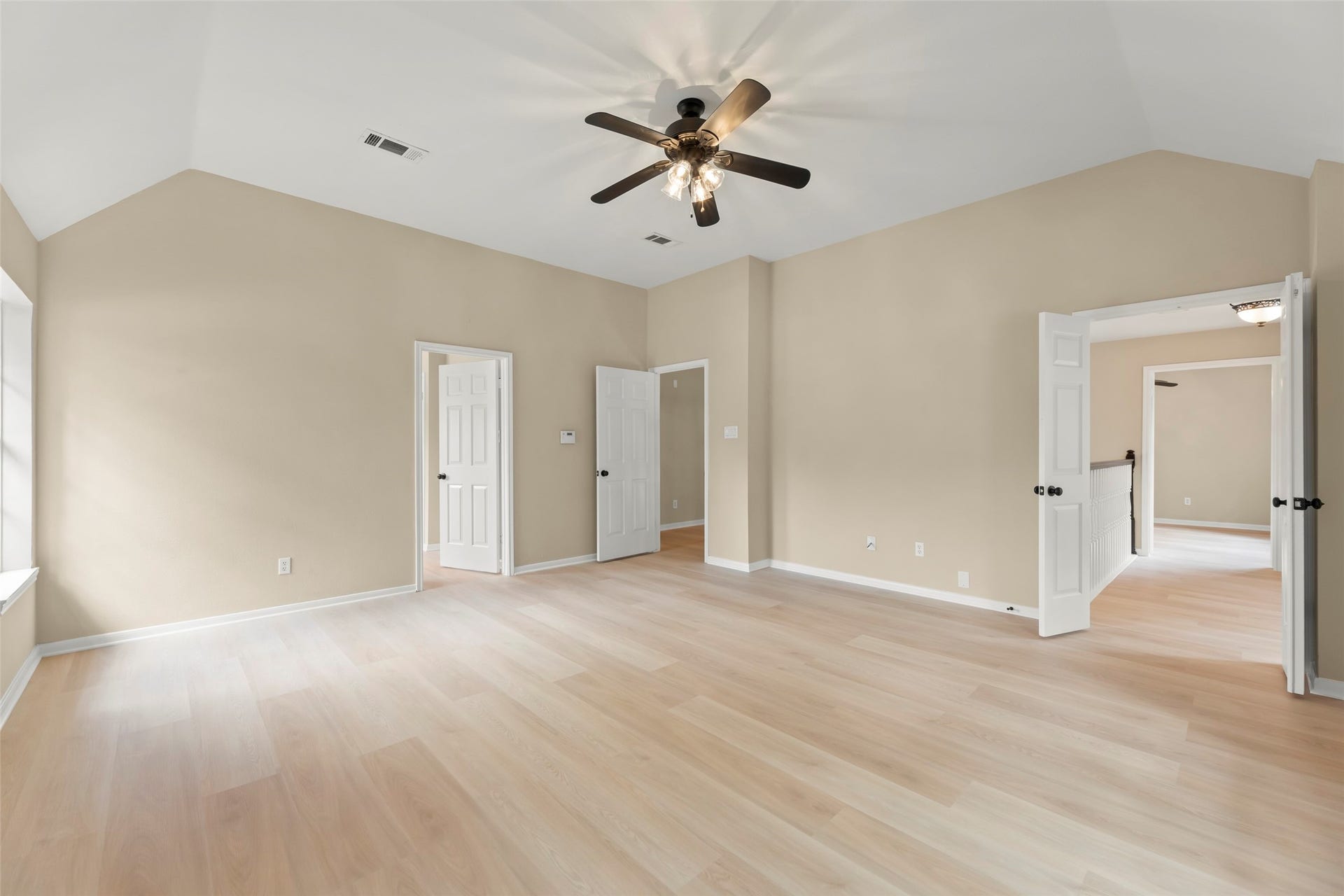 Primary bedroom with light wood flooring, neutral walls, and a ceiling fan. Features multiple doors leading to other areas, ample natural light, and high ceilings for an open feel.